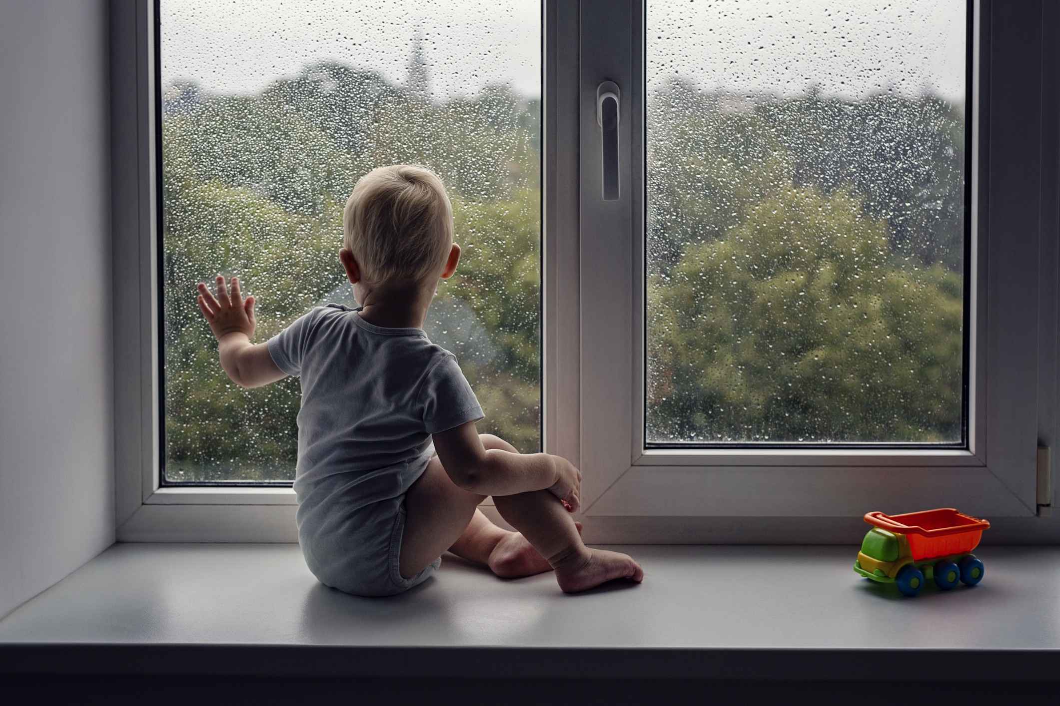 Infant boy sitting on a windowsill with his hand against the glass window with rain drops
